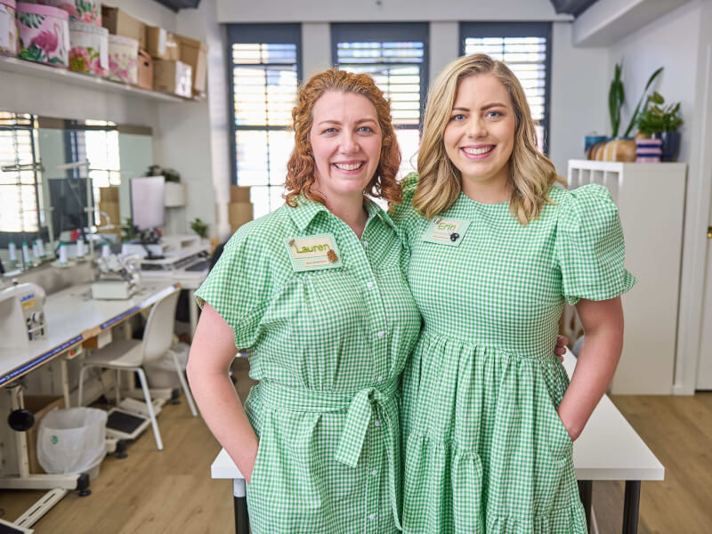 Two women in matching green gingham dresses are smiling at the camera
